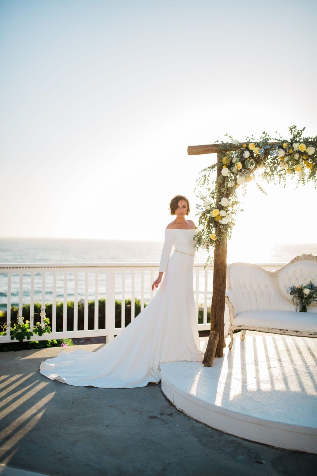 A bride in a white wedding dress standing in front of a wooden arch on the beach, making for a picturesque setting for Southern California weddings.