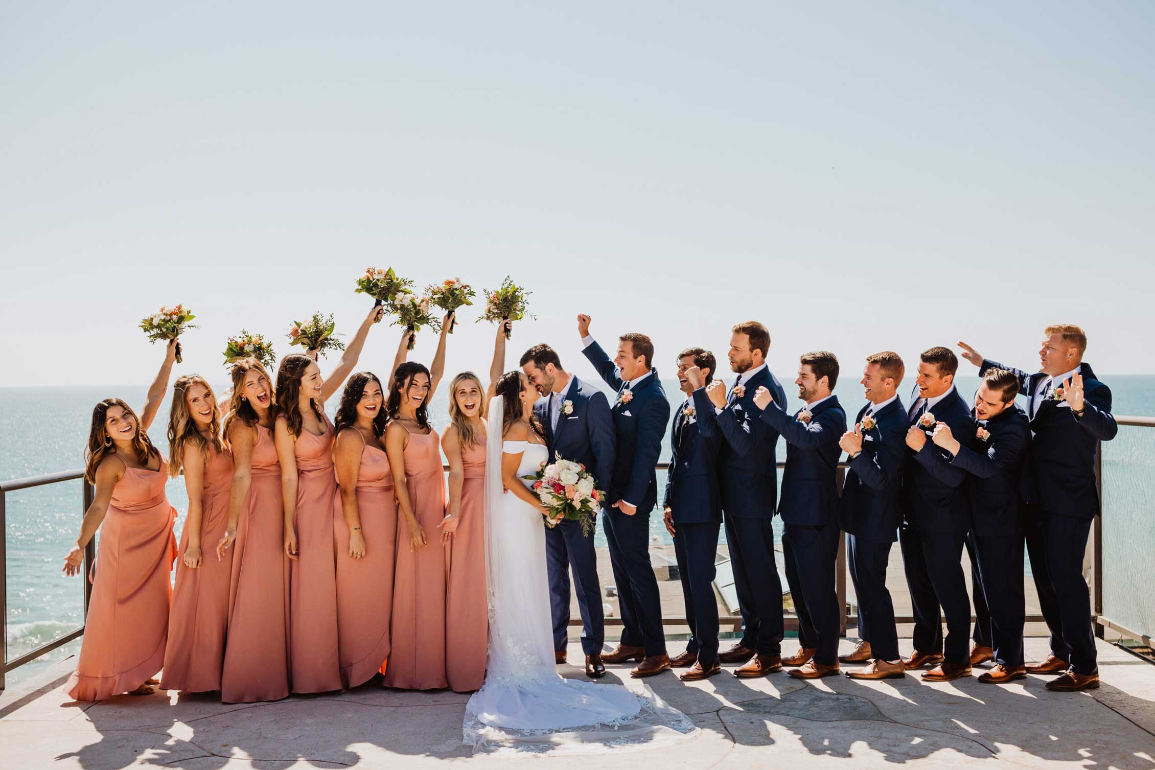 bride-and-groom-with-wedding-party-at-occasions-at-laguna-village Bride and groom kiss, surrounded by bridesmaids in pink dresses and groomsmen in blue suits, all celebrating on a sunny day by the sea.
