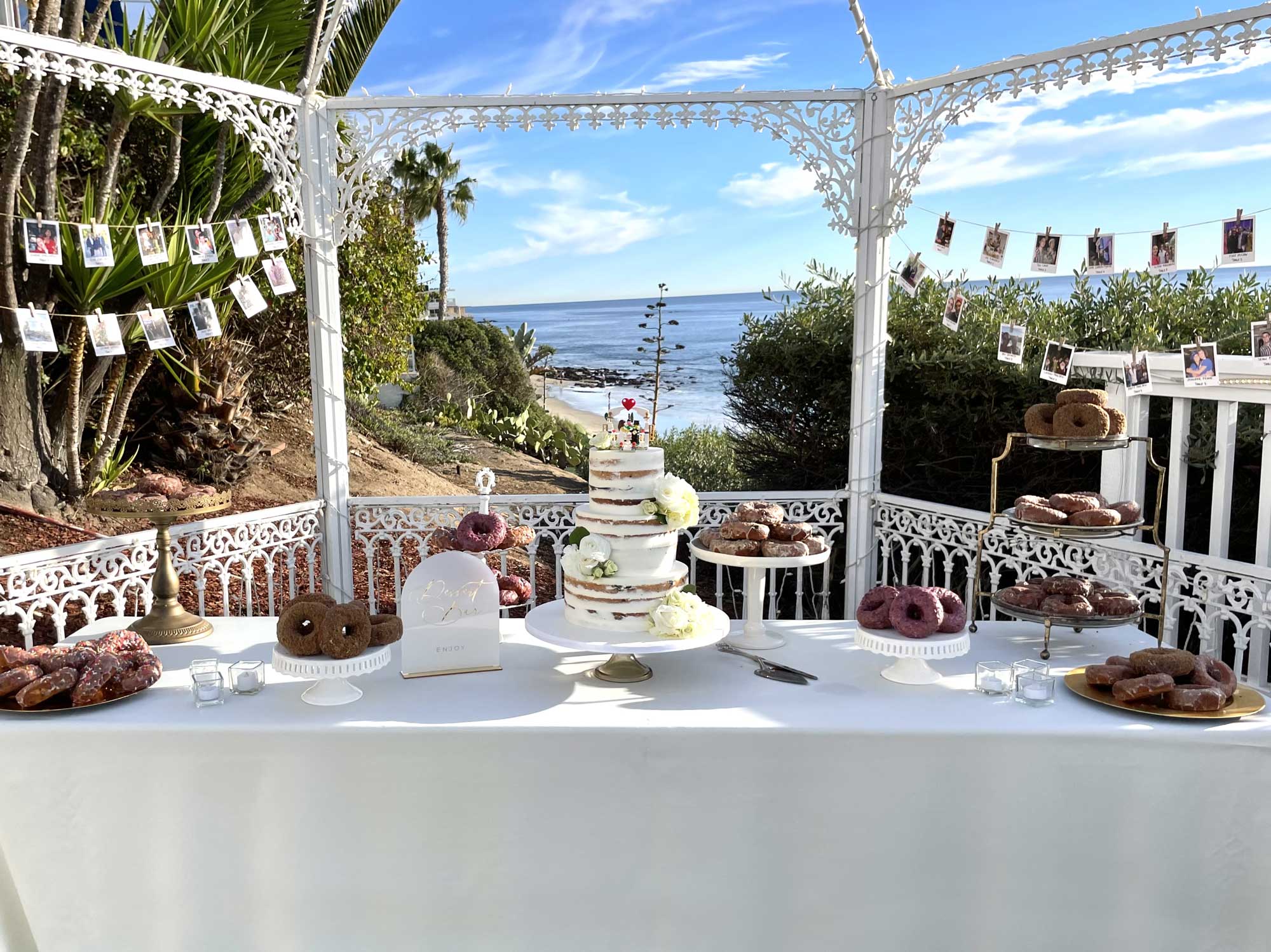 Wedding cake and assorted donuts displayed on a white table under a decorative gazebo overlooking the ocean. Photos are hung on a string in the background.